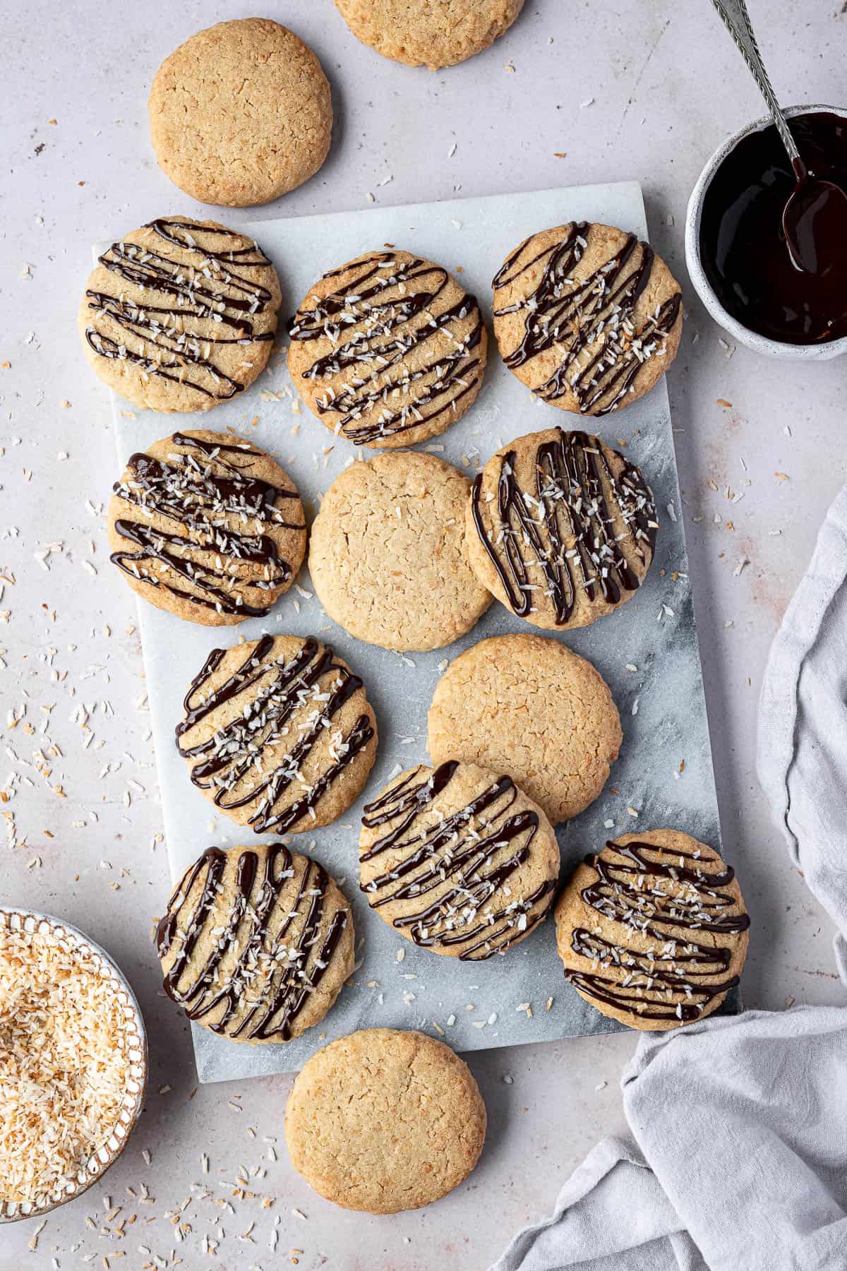 Vegan coconut cookies on a ,marble board with bowls of melted chocolate and toasted coconut.