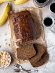 Banana gingerbread with cups of coffee, a tin of treacle, bananas and a bowl of candied ginger.