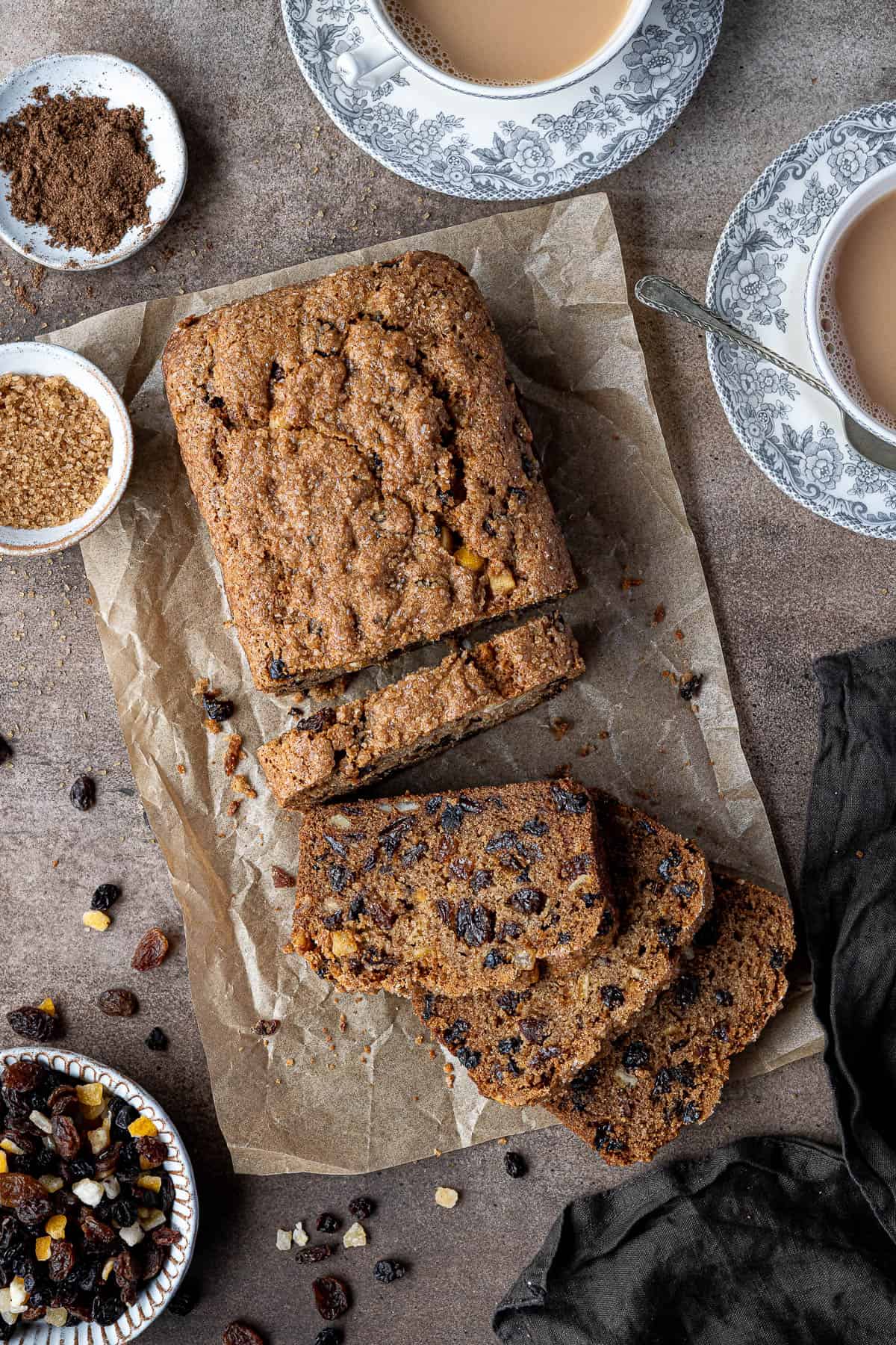 Vegan fruit cake loaf on a brown surface with cups of tea and bowls of fruit, sugar and spice.
