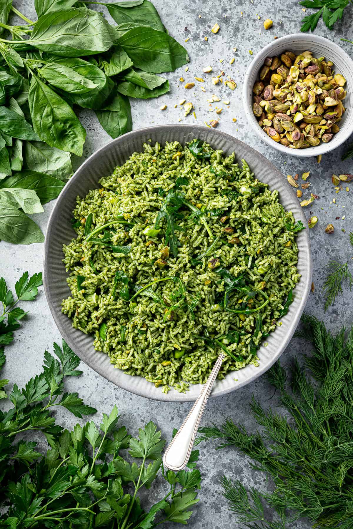 Herby rice in a grey bowl surrounded by fresh herbs and a bowl of pistachios.