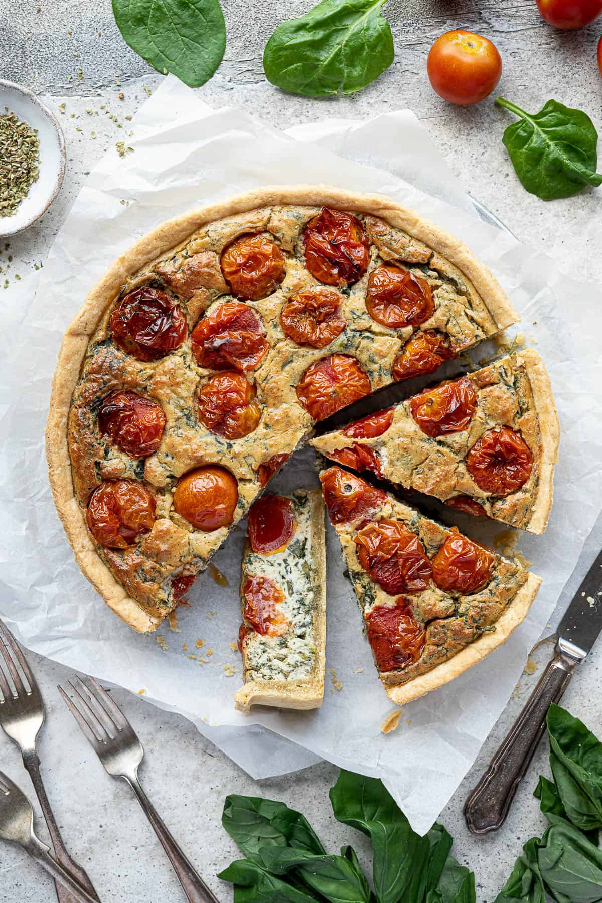 The sliced vegan tomato spinach quiche on a sheet of baking parchment surrounded by fresh basil, spinach and tomatoes.