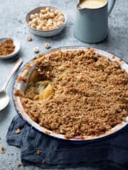 Rhubarb apple crumble in a round baking dish with a jug of custard and bowlds of hazelnuts and cinnamon.