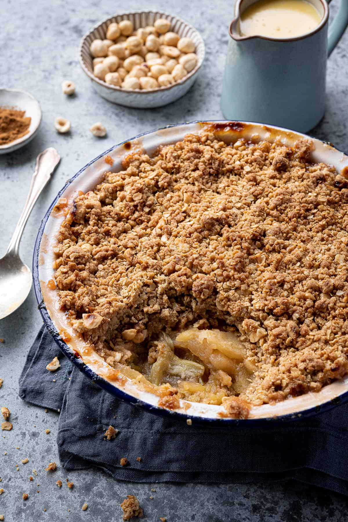 Close up of a dish of rhubarb apple crumble with a jug of custard and bowls of hazelnuts and cinnamon.