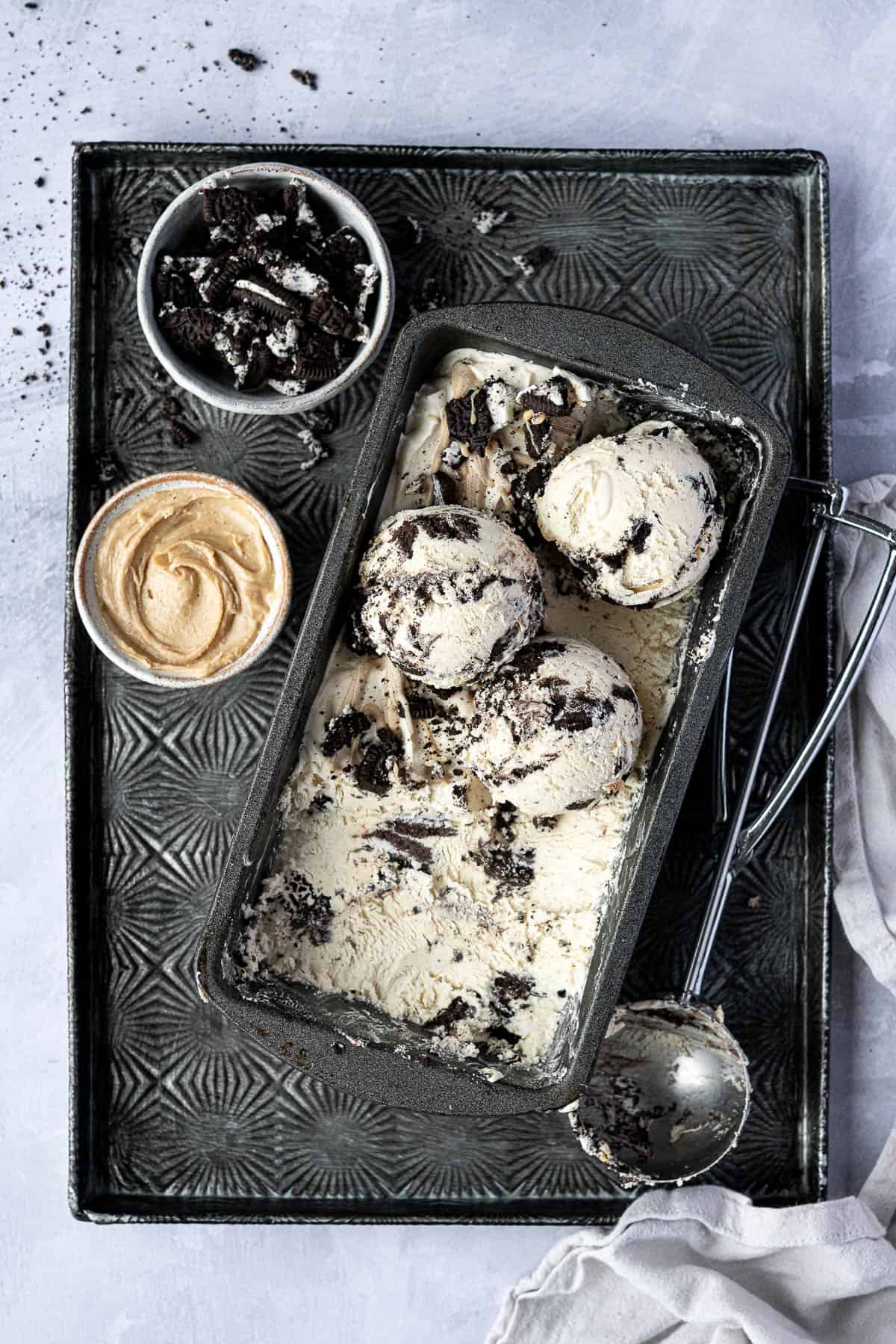 No-churn peanut butter Oreo ice cream in a loaf tin on a tray with an ice cream scoop and bowls of peanut butter and crushed Oreos.