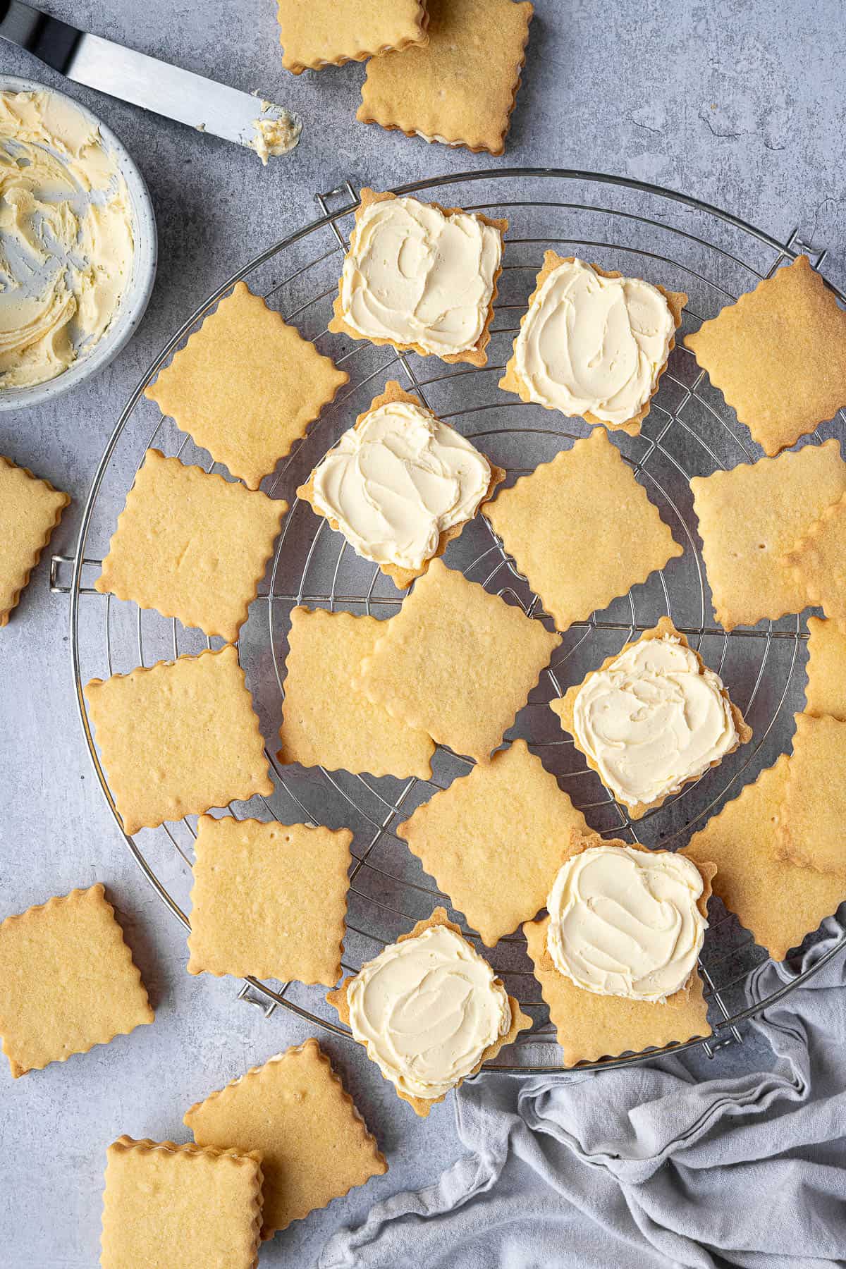 Vegan custard creams on a round wire cooling rack with a bowl of buttercream and a spatula.