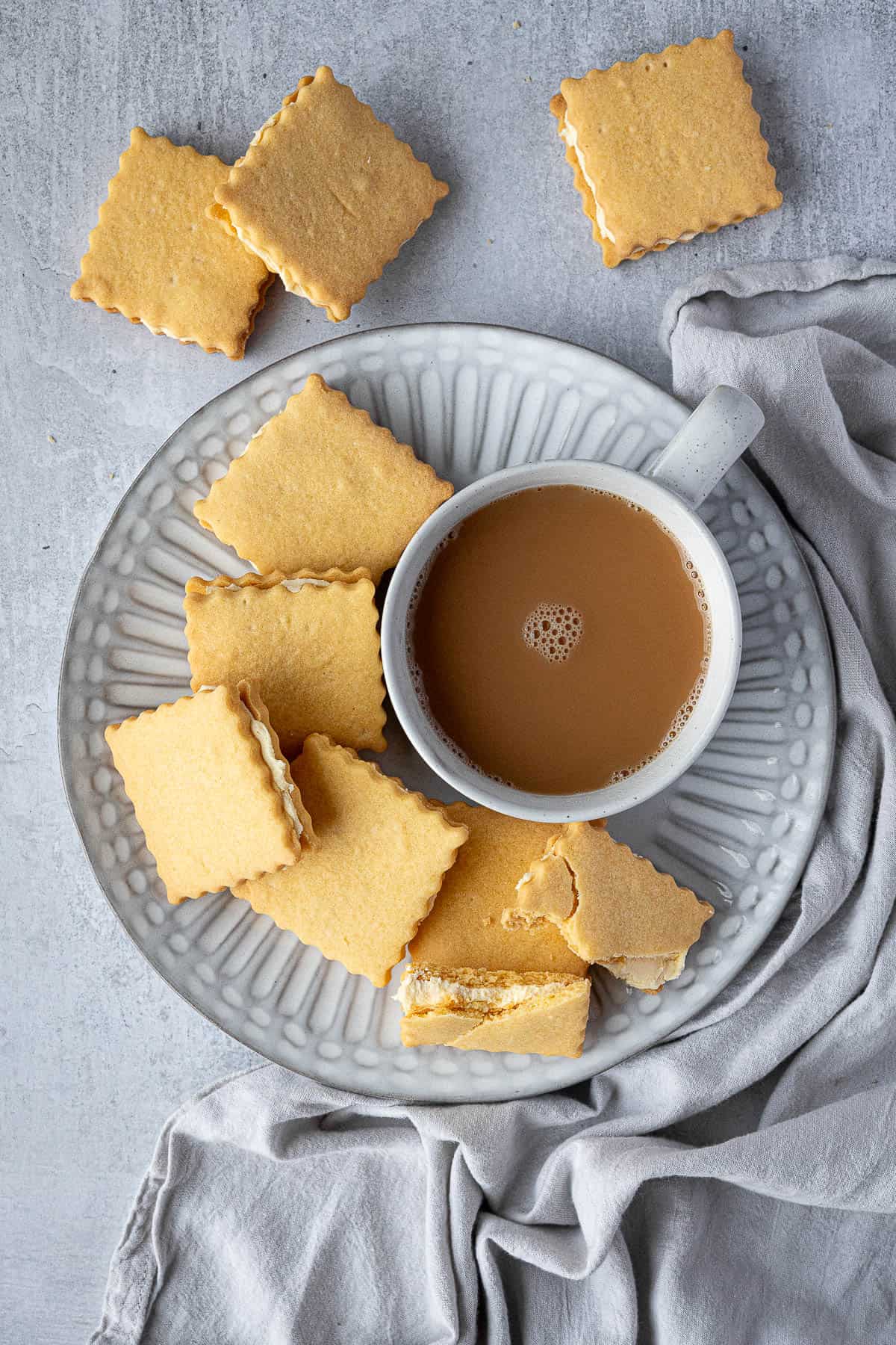 A white plate of custard cream cookies with a cup of tea.