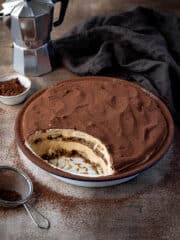 Pumpkin tiramisu in a round dish with a spoonful removed, a caffetiere, bowl of cocoa powder and small sieve in the background.
