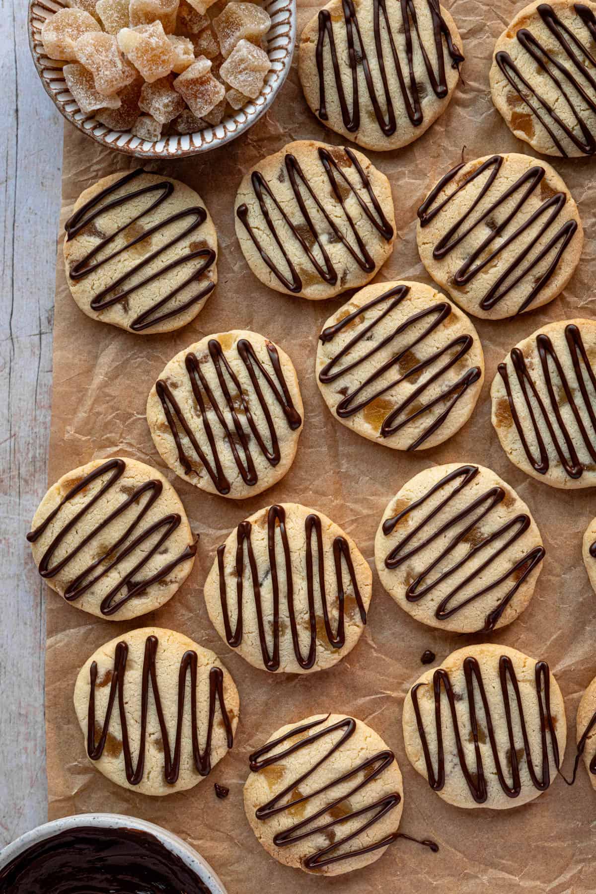 Ginger shortbread cookies on a sheet of brown baking parchment with bowls of candied ginger and chocolate.
