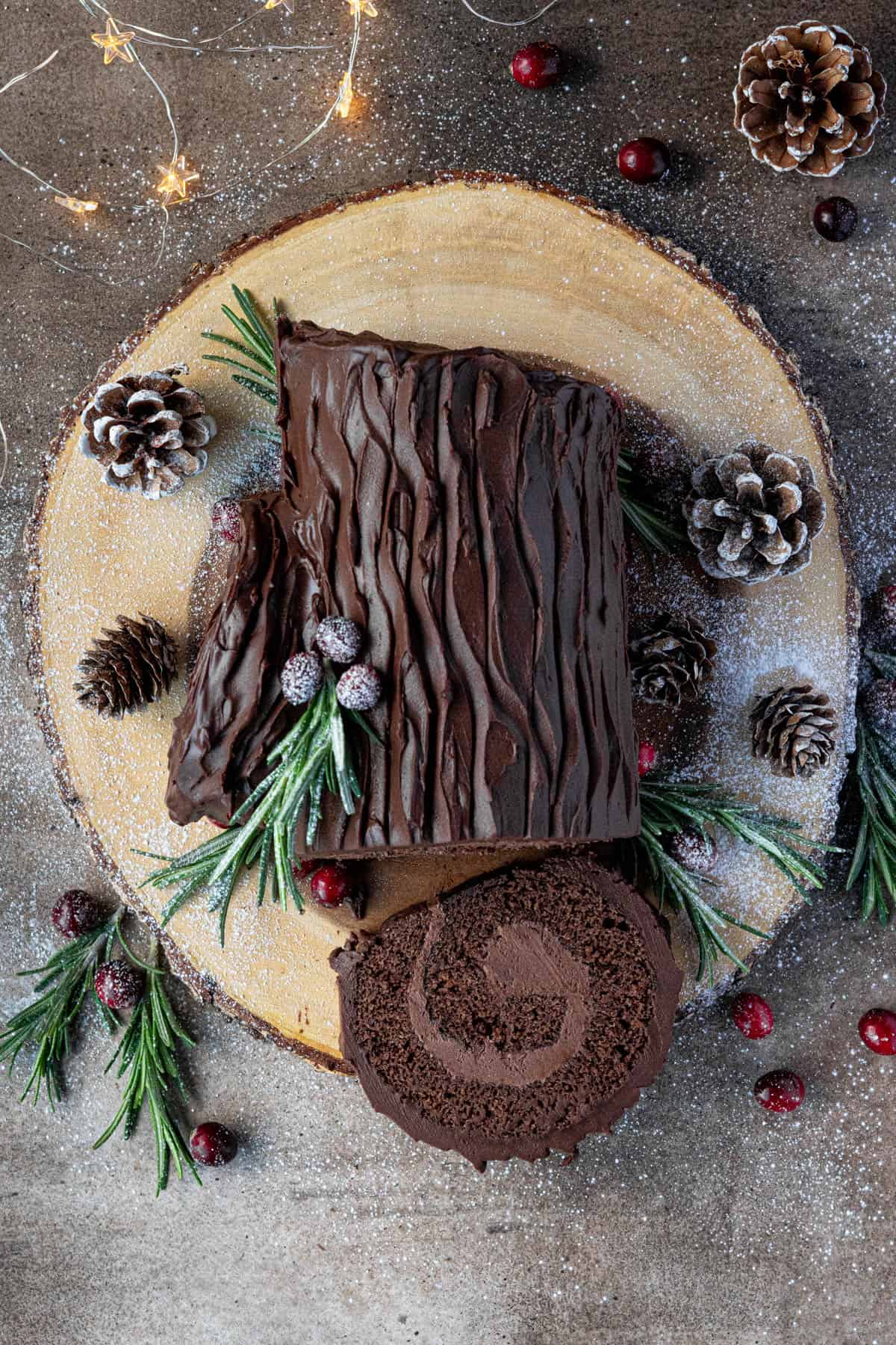 Top down shot of the sliced vegan yule log on a wooden board with pine cones and Christmas lights.