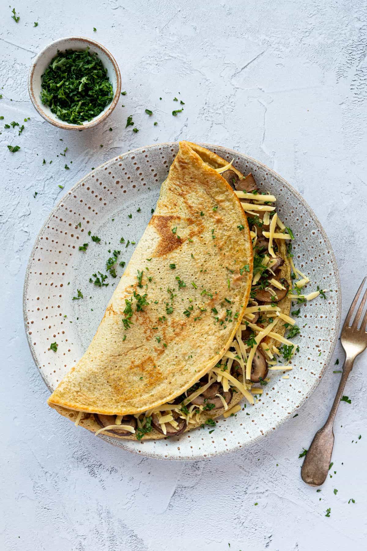 Savoury chickpea pancake filled with mushrooms and vegan cheese on a white plate with a bowl of chopped parsley.