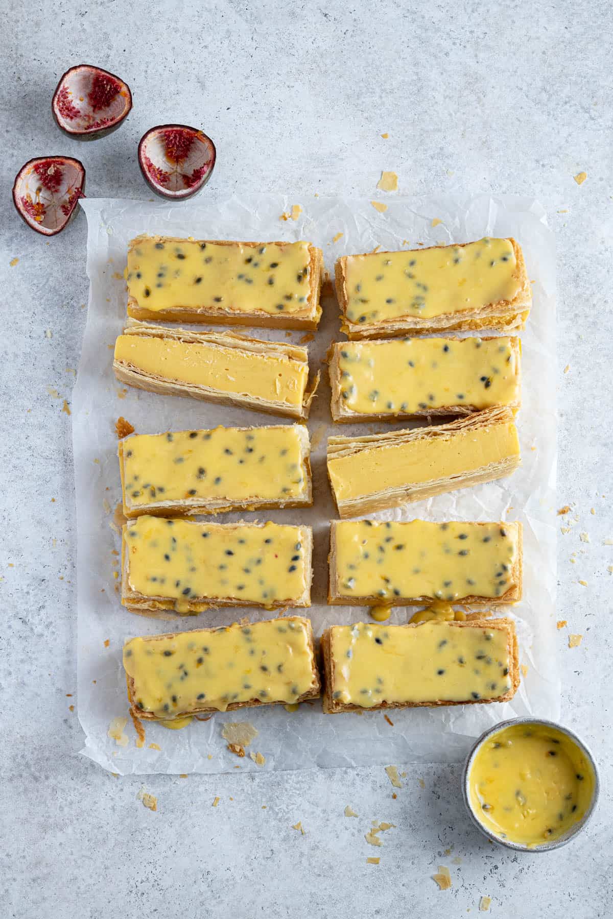 The sliced and glazed custard slices on a sheet of baking paper with passion fruit shells and a bowl of passion fruit glaze.