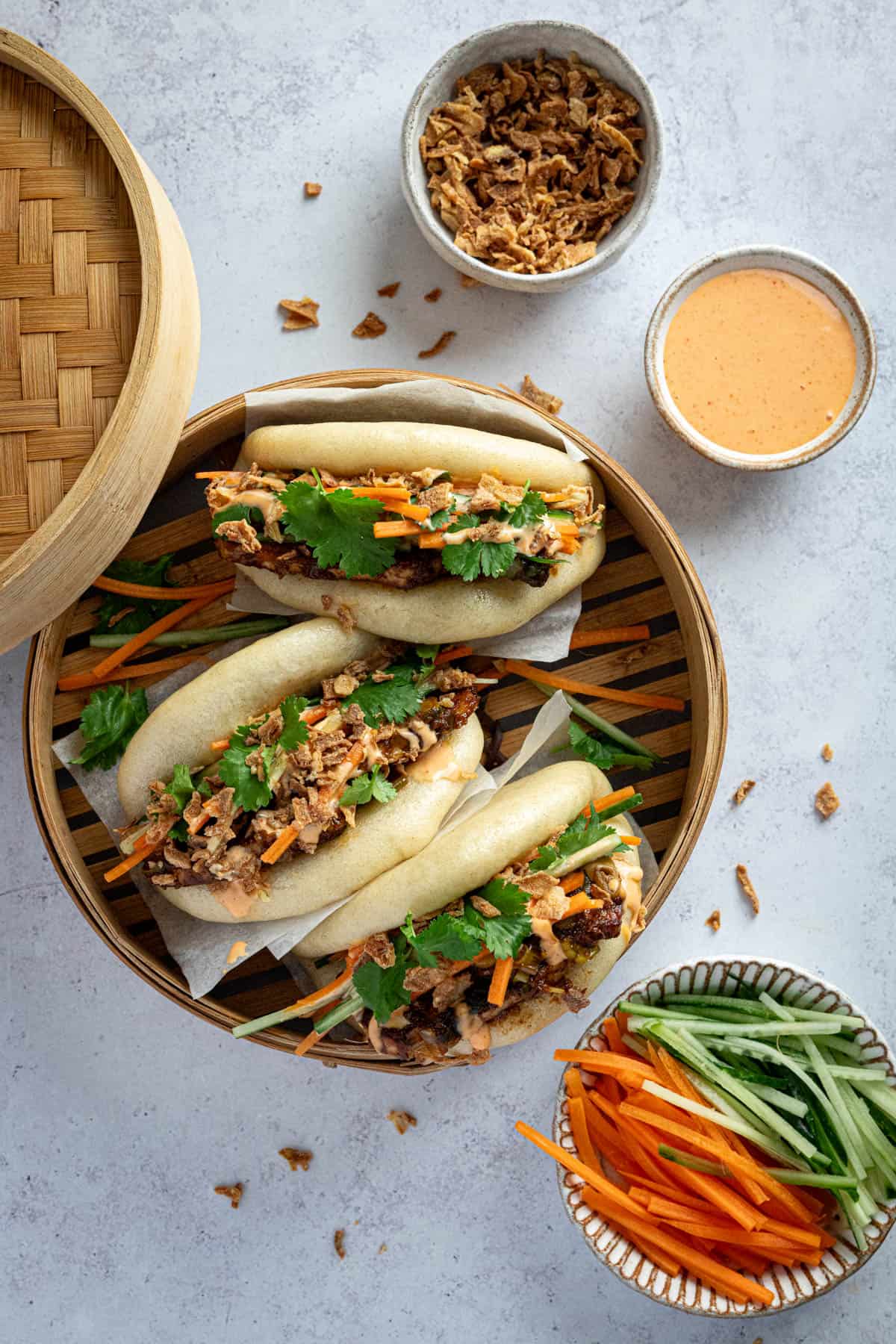 Three hoisin tofu bao buns in a bamboo steamer with bowls of ingredients.