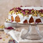 Vegan Persian love cake topped with pistchios and rose petals on a glass cake stand.