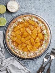 Caramelised pineapple coconut tart on a metal plate with forks, sliced limes and a bowl of desiccated coconut.