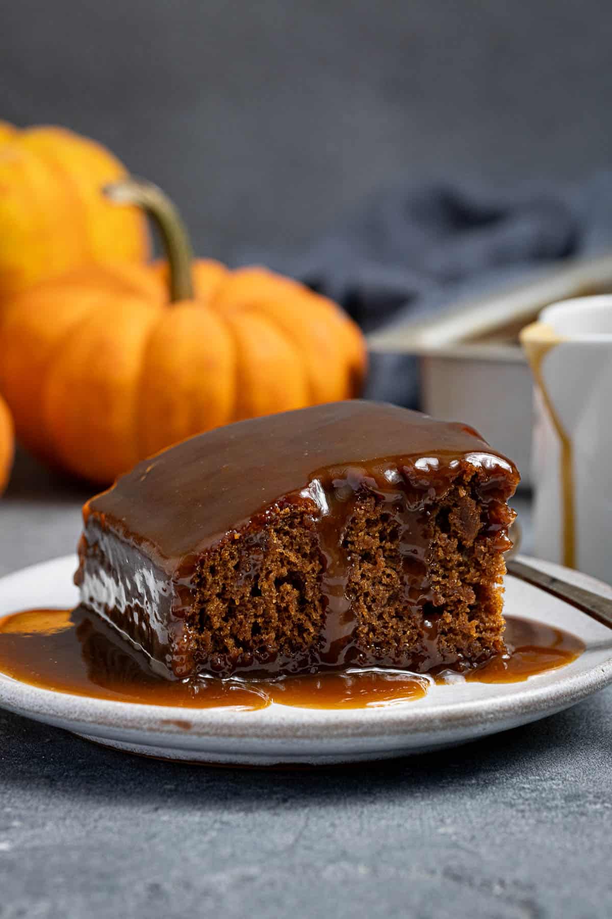 A slice of vegan pumpkin sticky toffee pudding on a white plate with mini pumpkins in the background.
