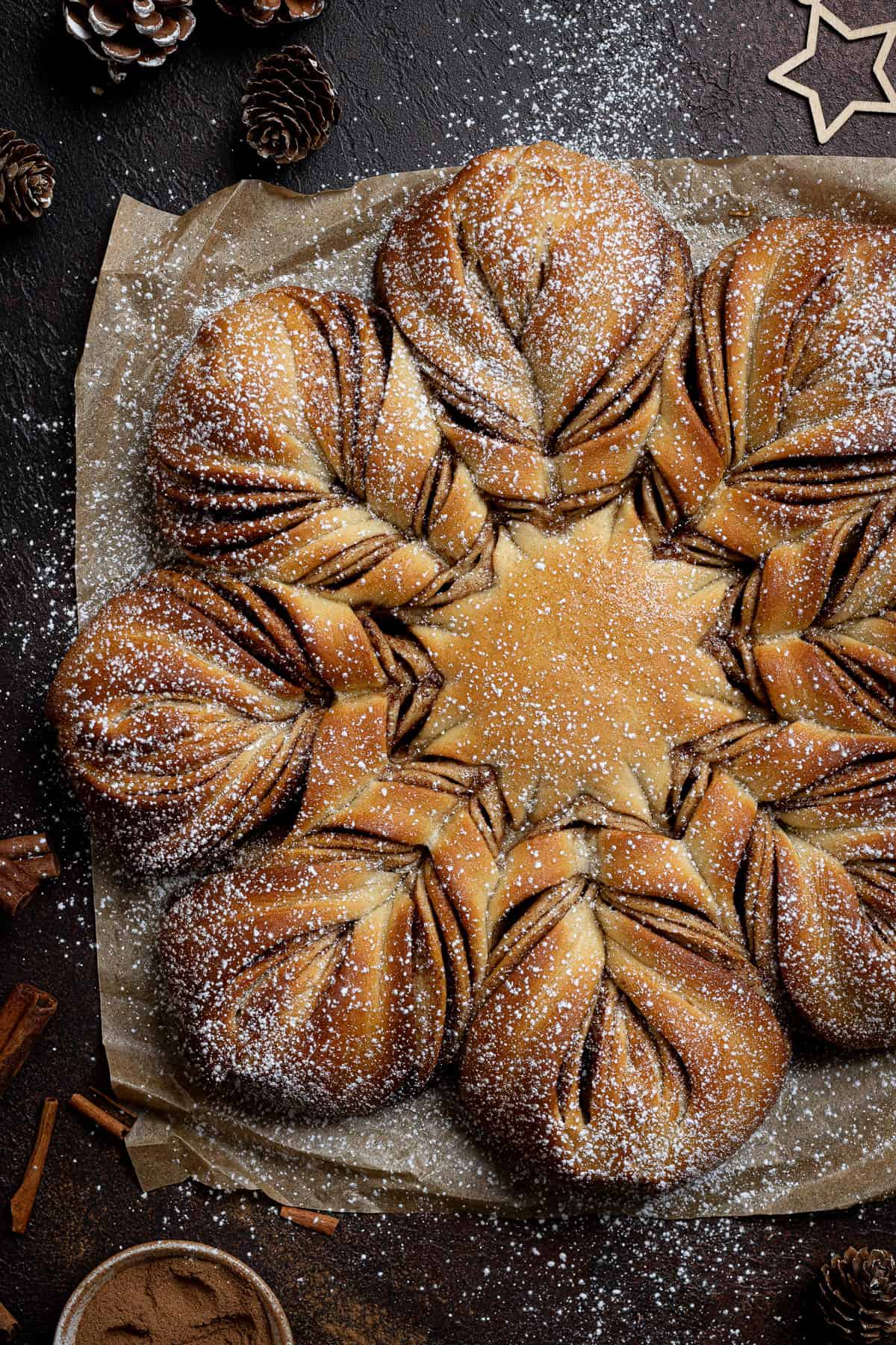 Vegan cinnamon star bread on a sheet of baking paper on a dark surface with a bowl of cinnamon, cinnamon sticks and pine cones.