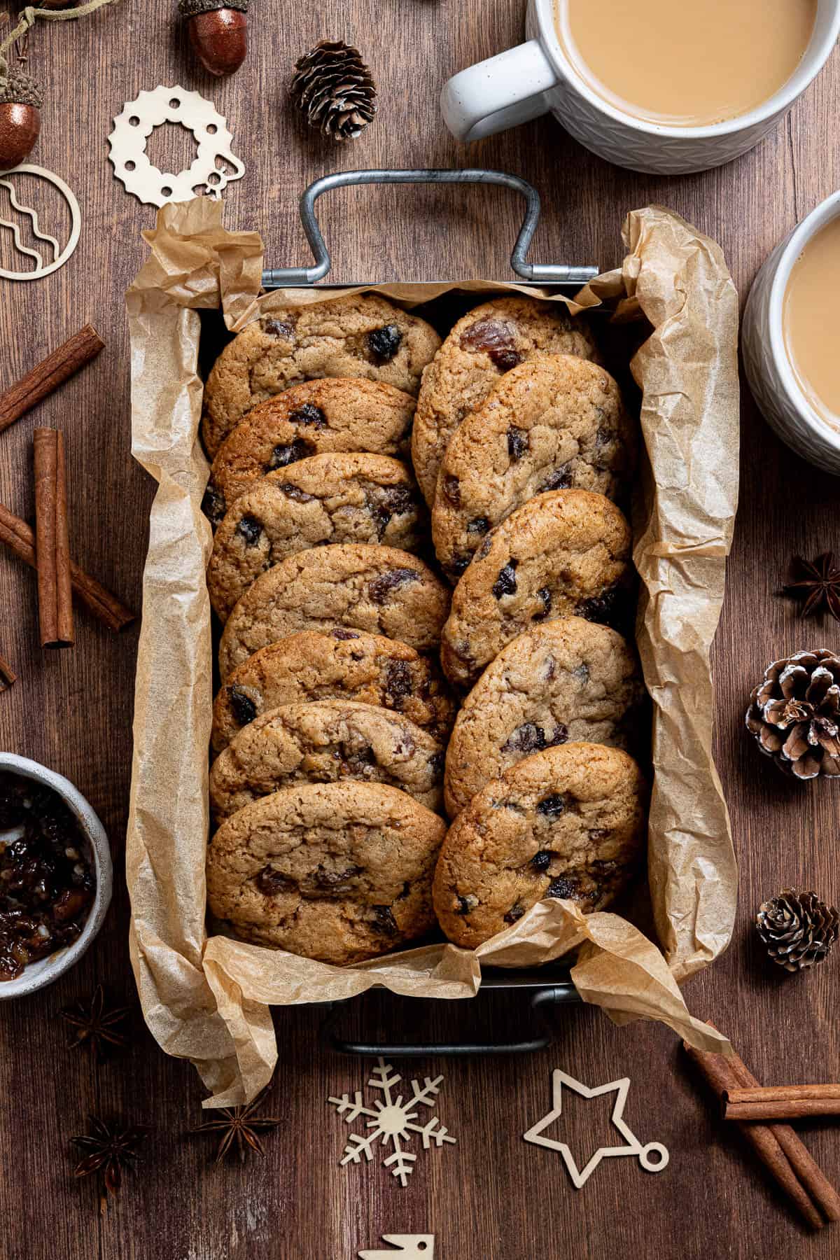 Vegan mincemeat cookies in a parchment lined box, surrounded by cups of tea, whole spices and Christmas decorations.