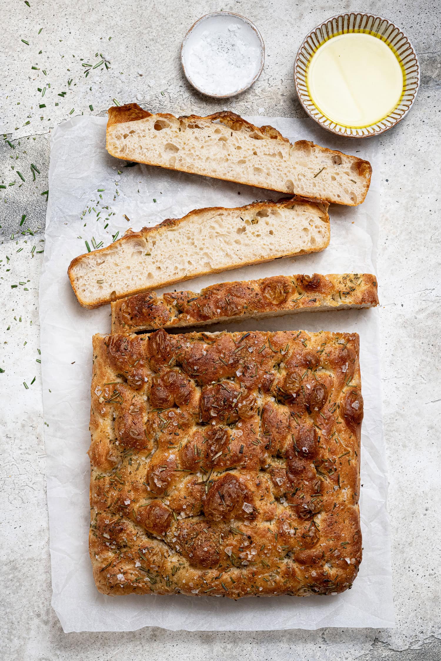 The sliced focaccia on a sheet of baking paper with bowls of olive oil and salt.