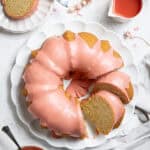 Flatlay of a vegan blood orange olive oil cake on a white plate with a bowl of white chocolate glaze, a jug of blood orange juice and dried flowers.