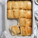 A pan of garlic Parker House rolls on a marble surface with a bowl of dried herbs and garlic cloves.