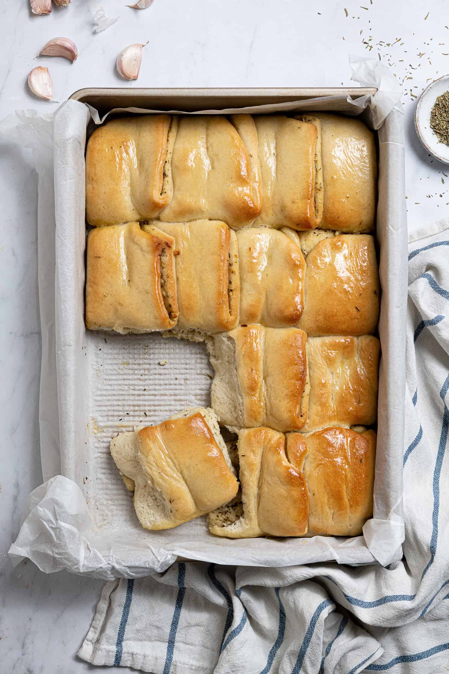 A pan of garlic Parker House rolls on a marble surface with a bowl of dried herbs and garlic cloves.