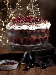 A glass bowl of vegan black forest trifle on a glass cake stand, with fairy lights in the background.