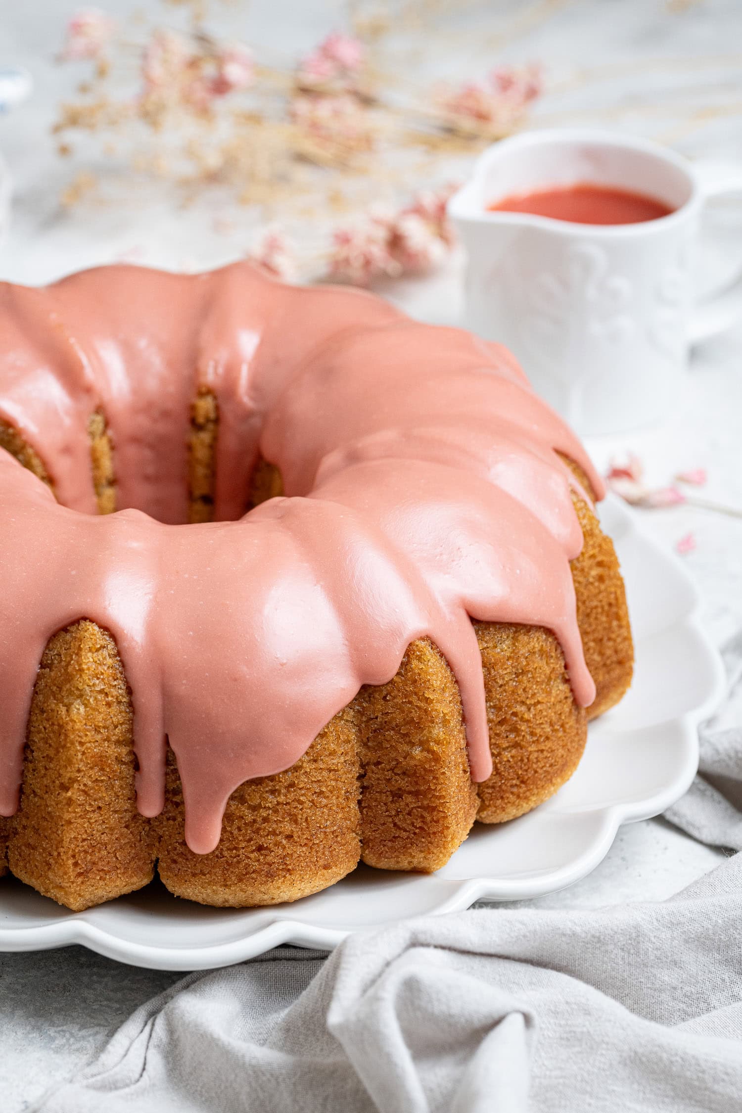 Vegan blood orange olive oil bundt cake on a white plate with a jug of blood orange juice and dried flowers in the background.