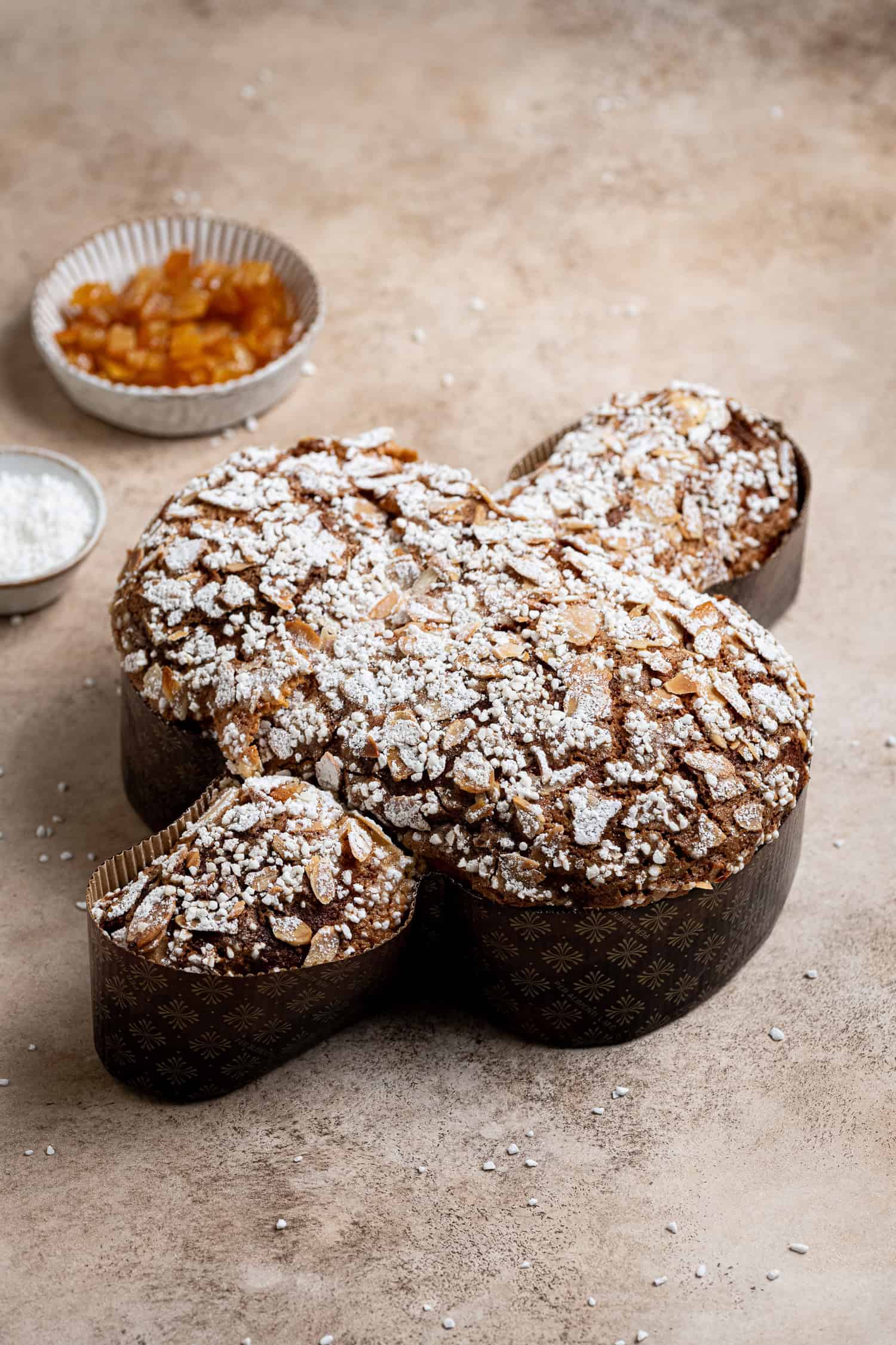 Vegan Colomba Pasquale on a brown surface with bowls of candied peel and pearl sugar.