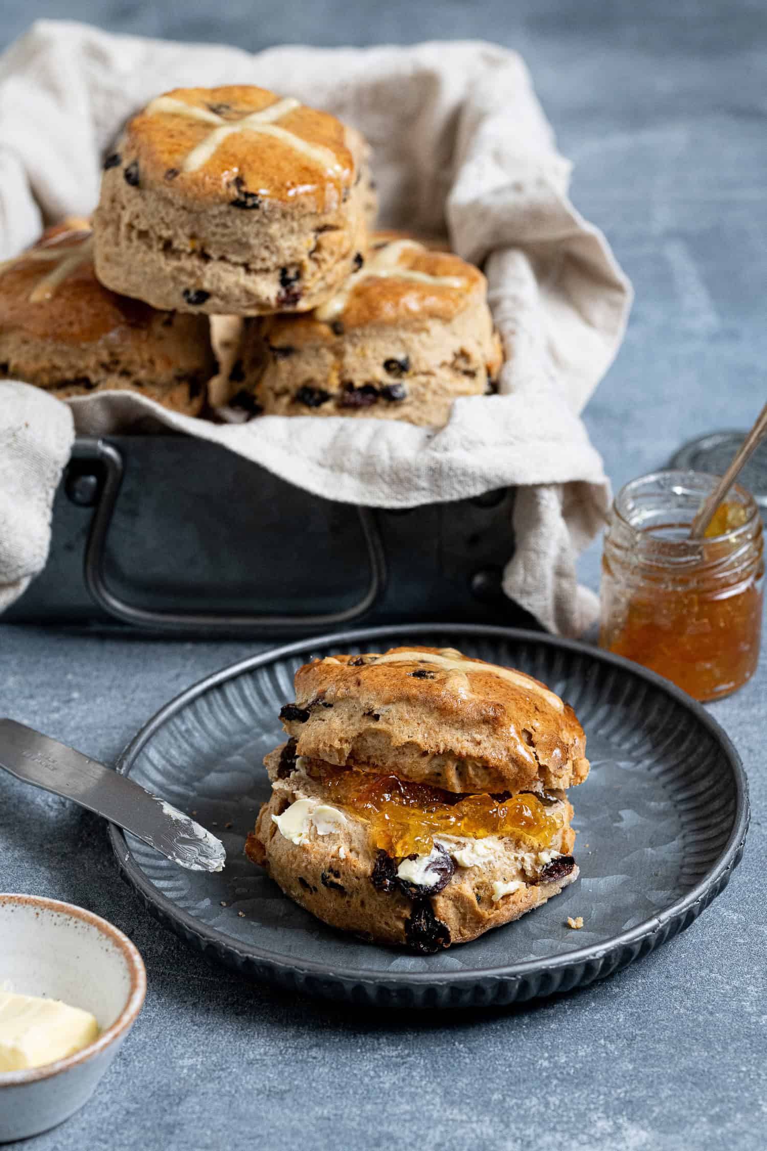 A scone sliced in half and filled with butter and marmalade on a metal plate with a box full of scones in the background.