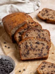 Sliced vegan chocolate chip banana bread on a sheet of baking paper with a bowl of chocolate chips.
