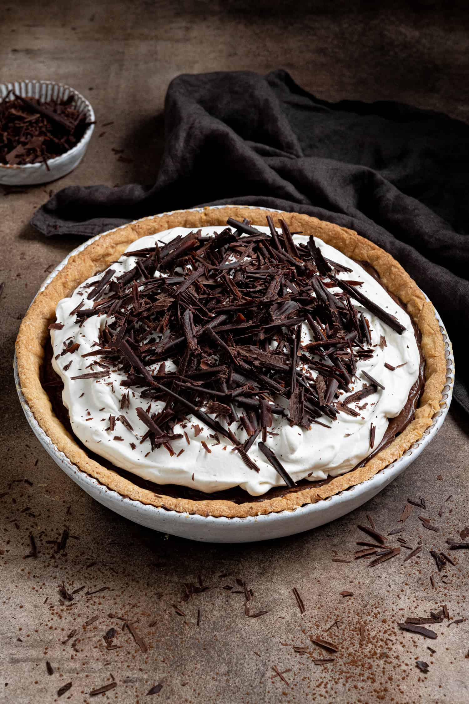 Vegan chocolate cream pie on a brown surface with a bowl of chocolate shavings and a brown cloth.