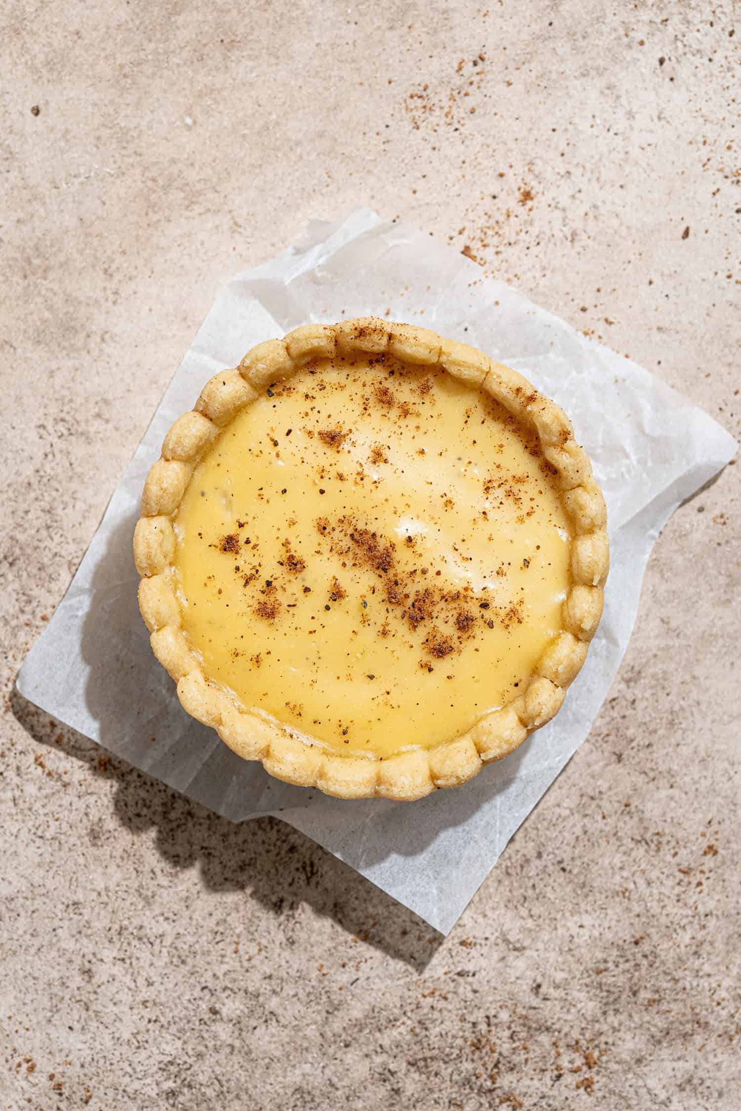 A vegan custard tart on a square of baking paper on a brown surface.