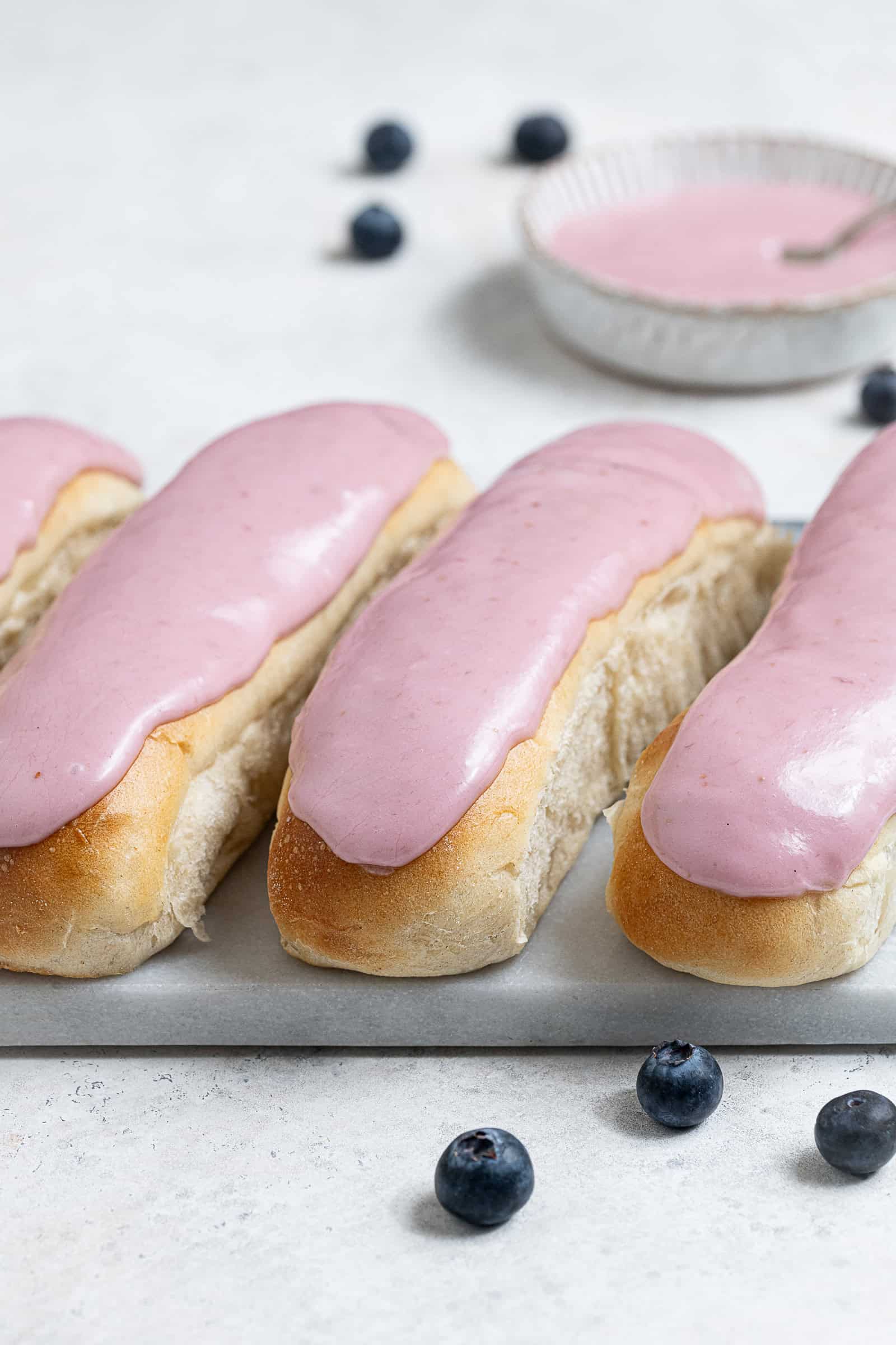 A row of vegan iced buns on a marble board with a bowl of pink icing.