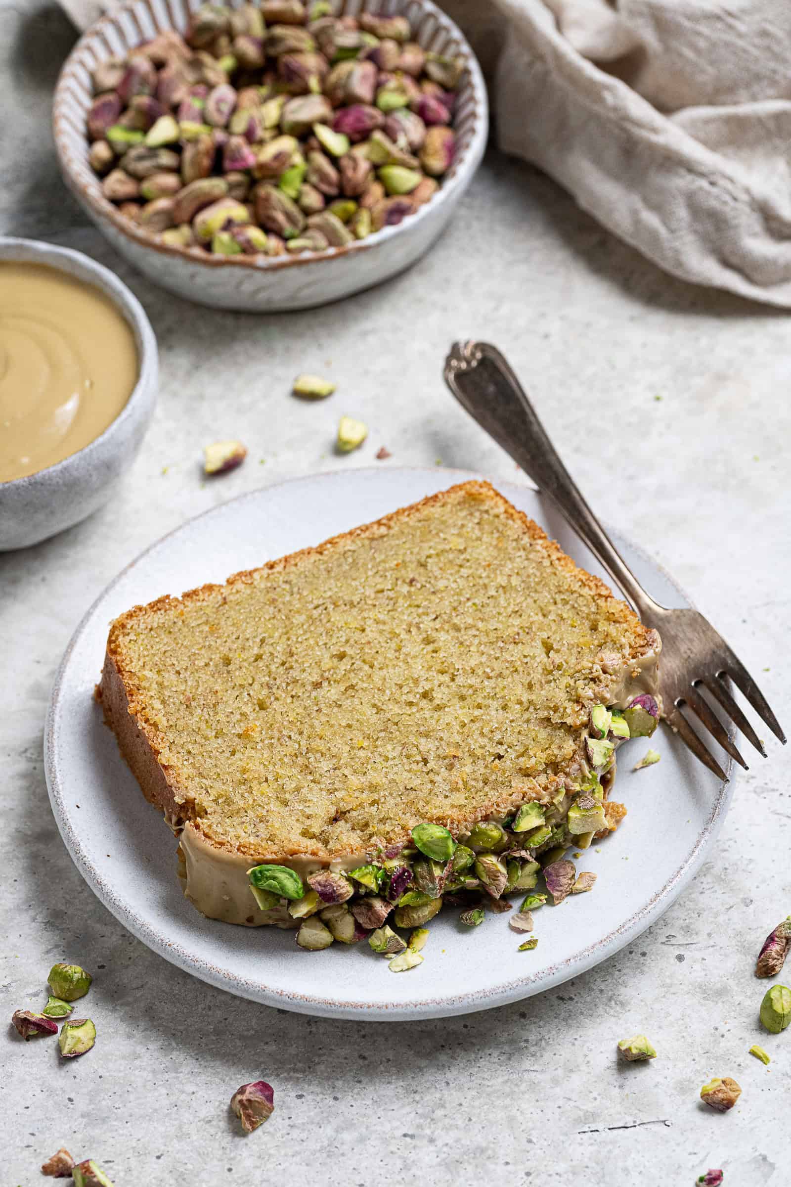A slice of vegan pistachio loaf cake on a white plate with bowls of pistachios and pistachio cream.