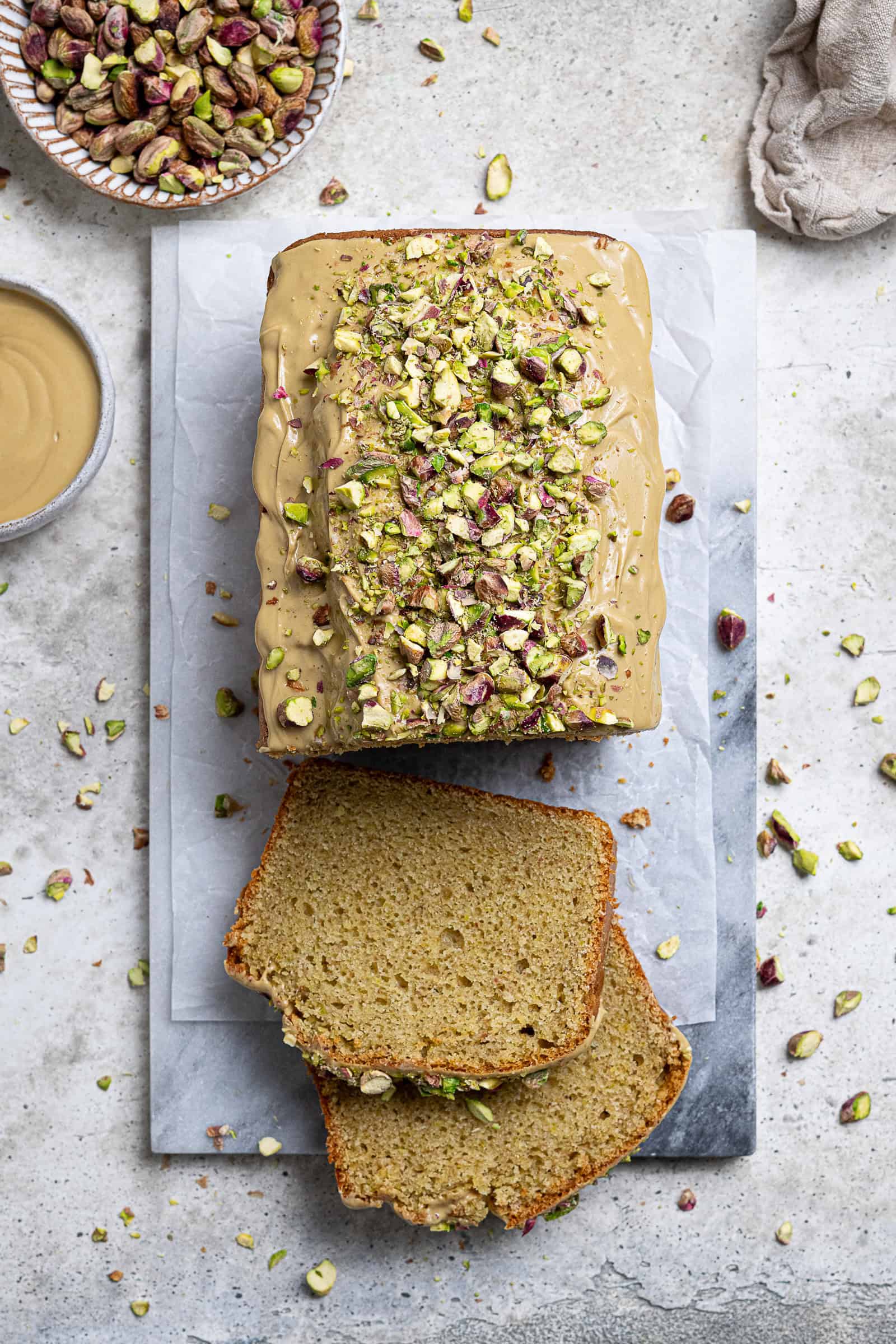 Flatlay of vegan pistachio loaf cake on a marble board with two slices cut and bowls of pistachios and pistachio cream in the background.