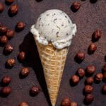 An ice cream cone with a scoop of vegan hazelnut stracciatella ice cream on a dark surface surrounded by hazelnuts.