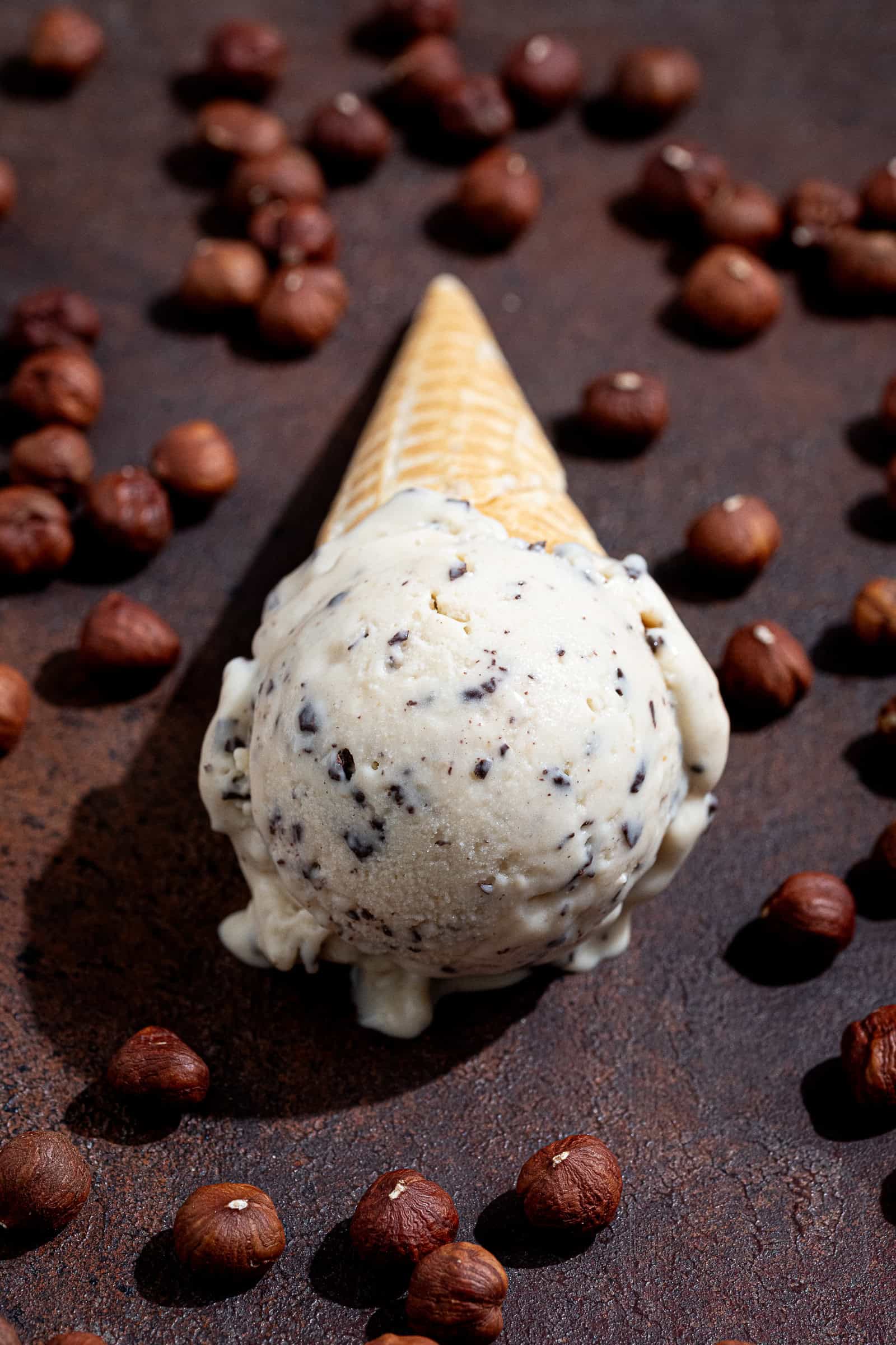 Close up shot of a scoop of vegan hazelnut stracciatella ice cream in an ice cream cone, surrounded by hazelnuts.
