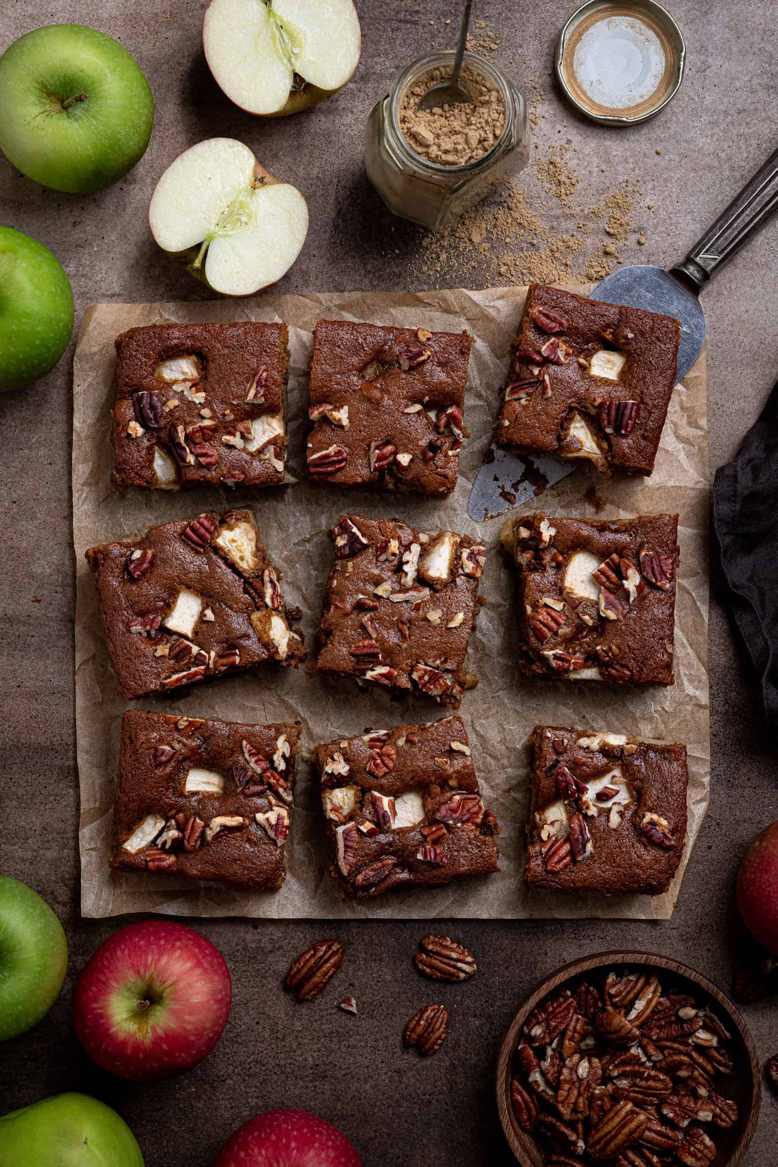 Flatlay of squares of vegan apple gingerbread cake on a dark surface surrounded by apples, pecans and a jar of ginger.