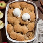 A baking dish of vegan apple cobbler on a wooden table surrounded by fresh apples and cinnamon.