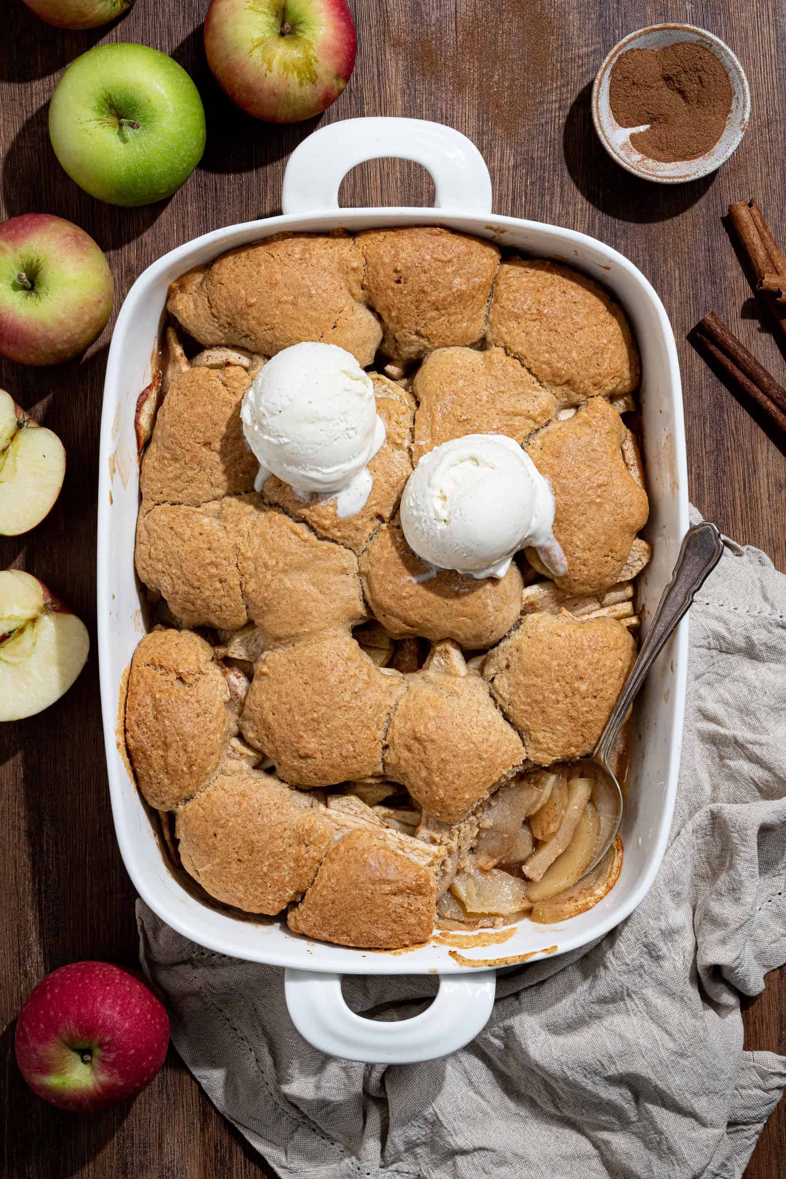 A baking dish of vegan apple cobbler on a wooden table surrounded by fresh apples and cinnamon.