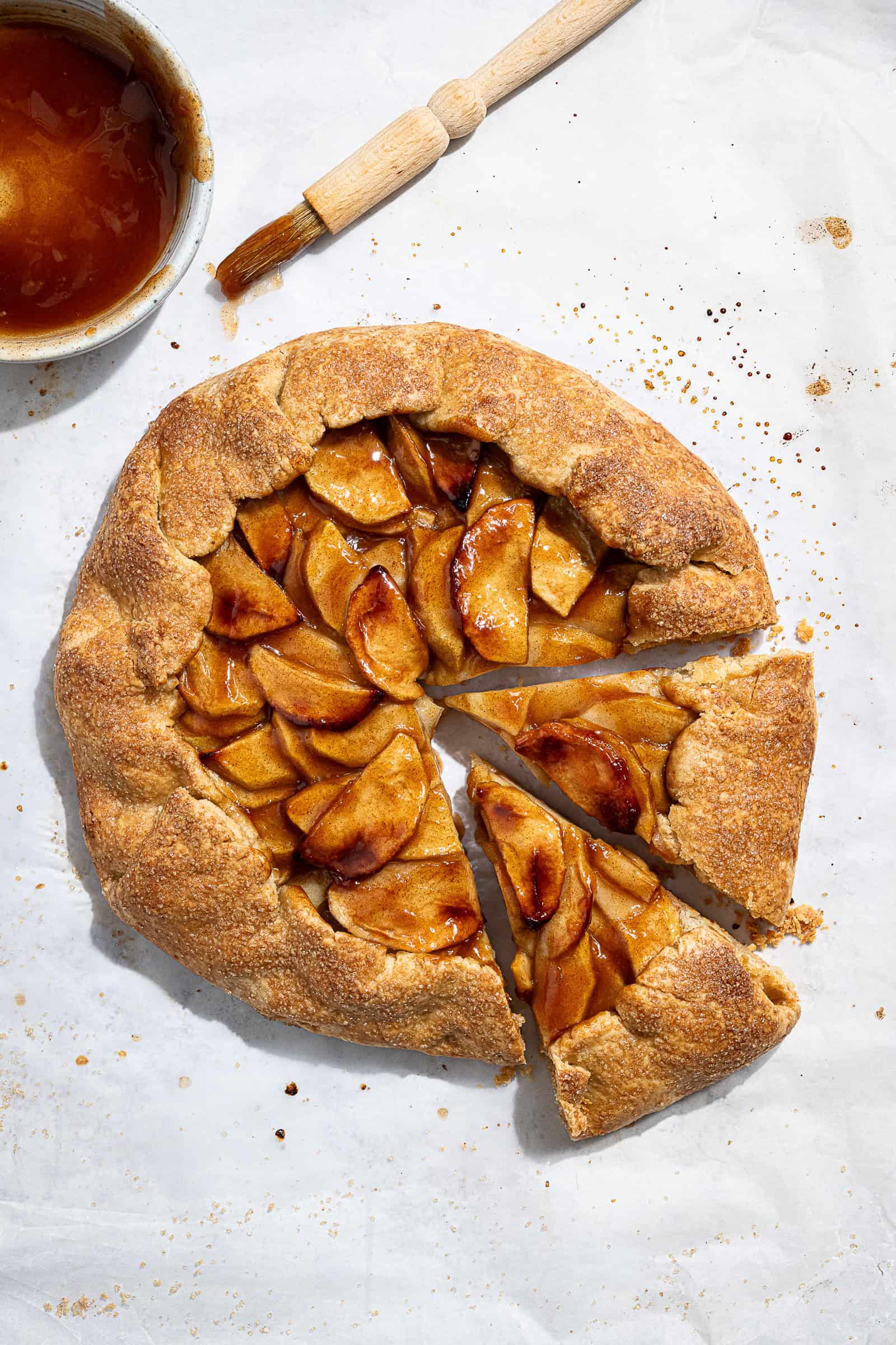 Vegan apple galette with two slices cut on a sheet of baking paper with a bowl of glaze and a pastry brush.