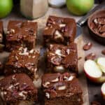Squares of vegan apple gingerbread cake on a sheet of baking paper with fresh apples, pecans and a jar of ginger.
