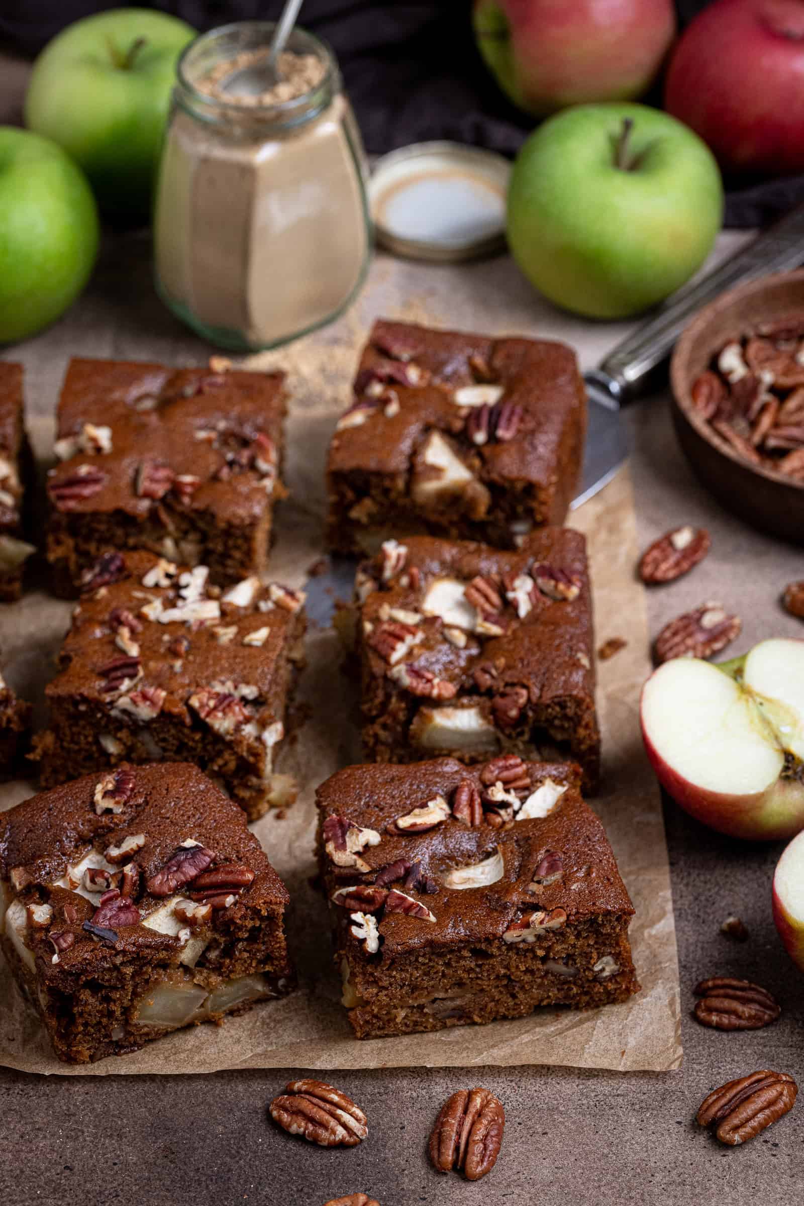 Squares of vegan apple gingerbread cake on a sheet of baking paper with fresh apples, pecans and a jar of ginger.