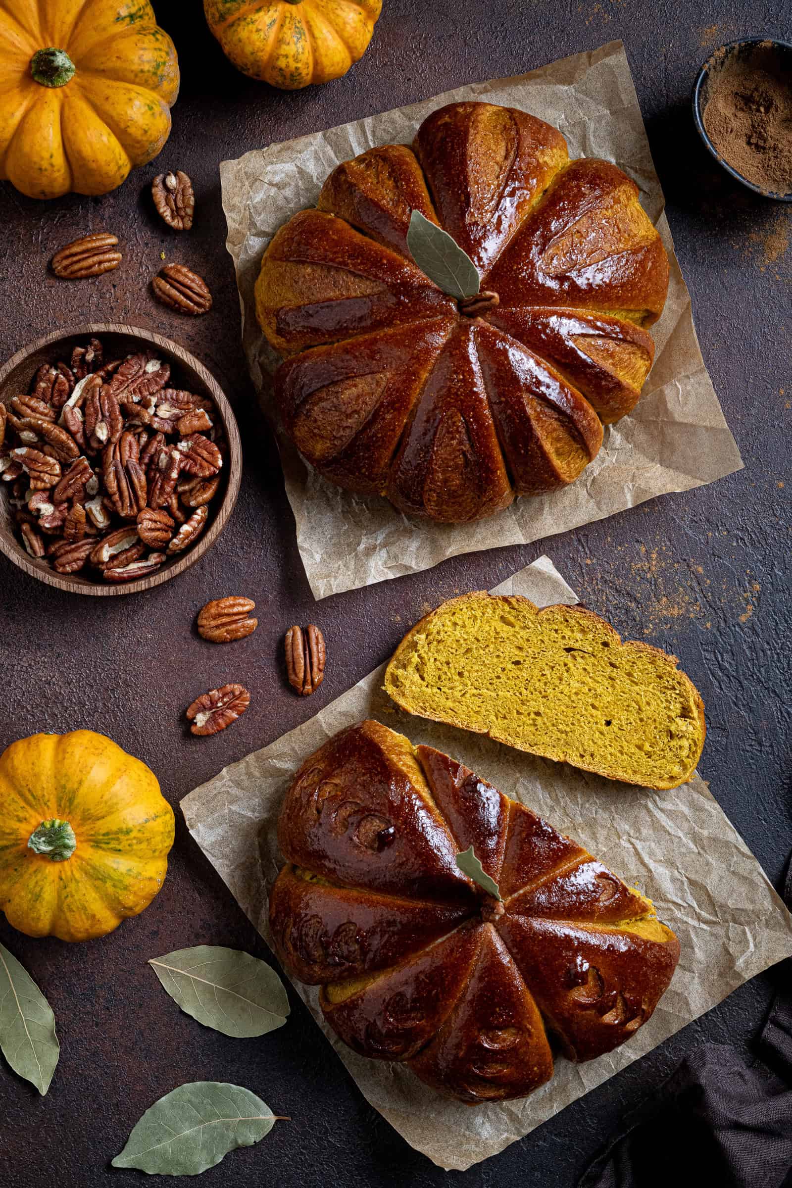 Flatlay of two loaves of pumpkin shaped pumpkin bread, one with a slice cut, surrounded by mini pumpkins, pecan nuts and bay leaves.