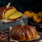 Two loaves of pumpkin shaped pumpkin bread on a dark surface, with mini pumpkins and a bowl of pecan nuts in the background.