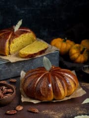 Two loaves of pumpkin shaped pumpkin bread on a dark surface, with mini pumpkins and a bowl of pecan nuts in the background.