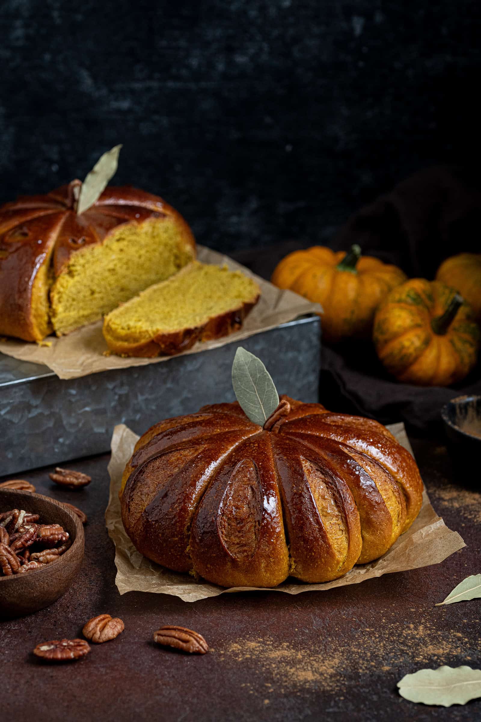 Two loaves of pumpkin shaped pumpkin bread on a dark surface, with mini pumpkins and a bowl of pecan nuts in the background.