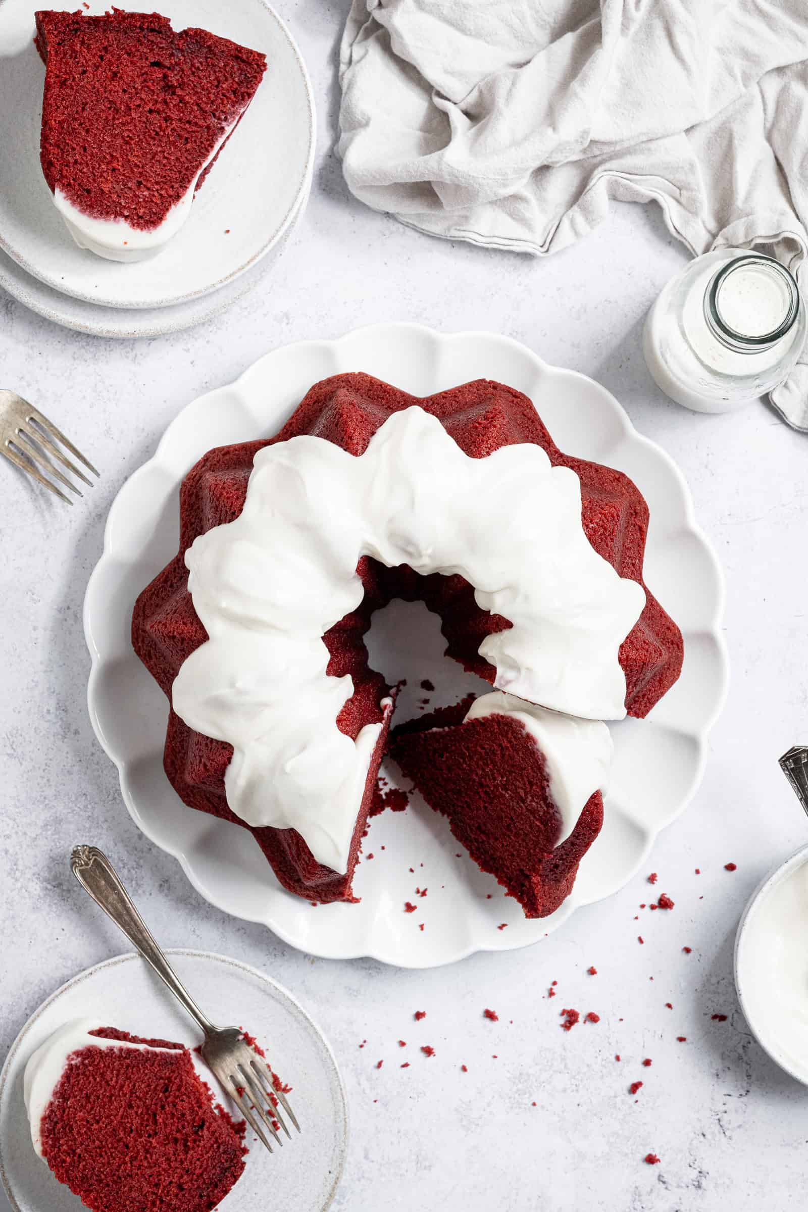 Flatlay of vegan red velvet bundt cake on a white plate, with two small plates with slices of cake and a glass bottle of milk.