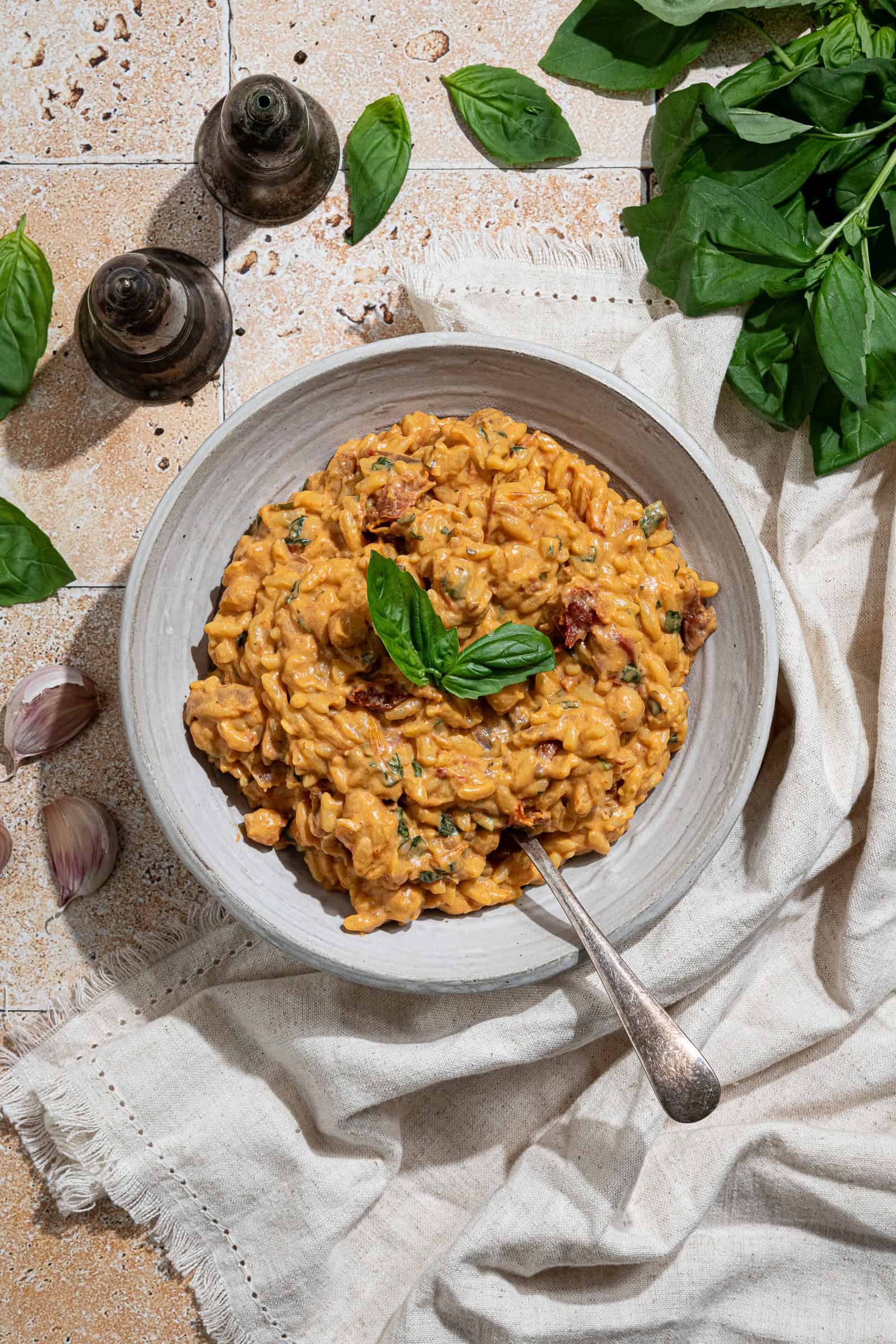Creamy vegan marry me orzo in a white bowl with a spoon on a tiled surface with fresh basil and salt and pepper shakers.