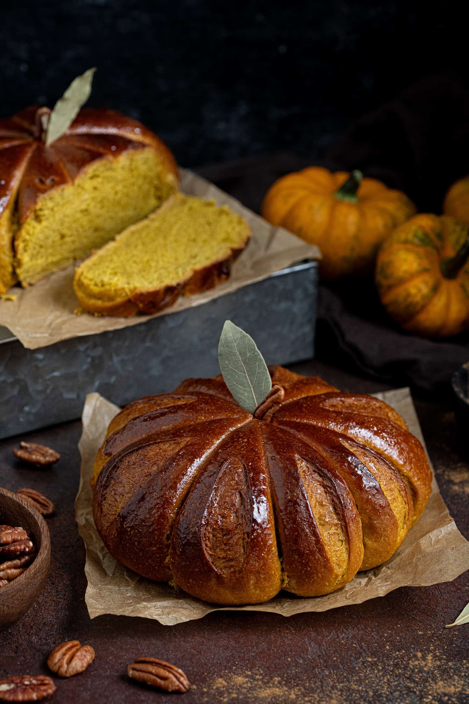 A loaf of pumpkin shaped bread on a dark surface, with another sliced loaf on top of a metal box in the background.